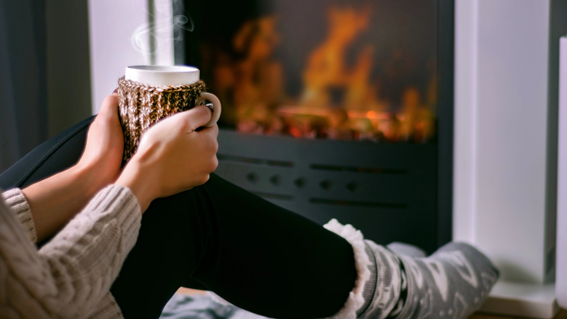 a woman holding a warm cup of tea in front of a fireplace